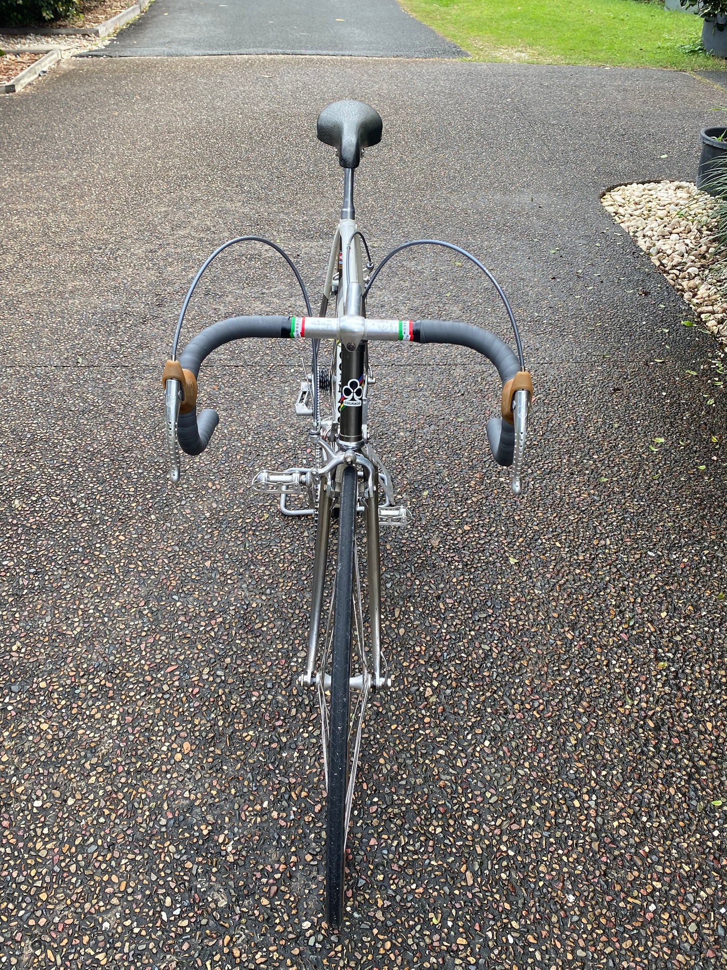 A vintage road bicycle with a silver frame and black drop handlebars is parked on a gravel driveway.