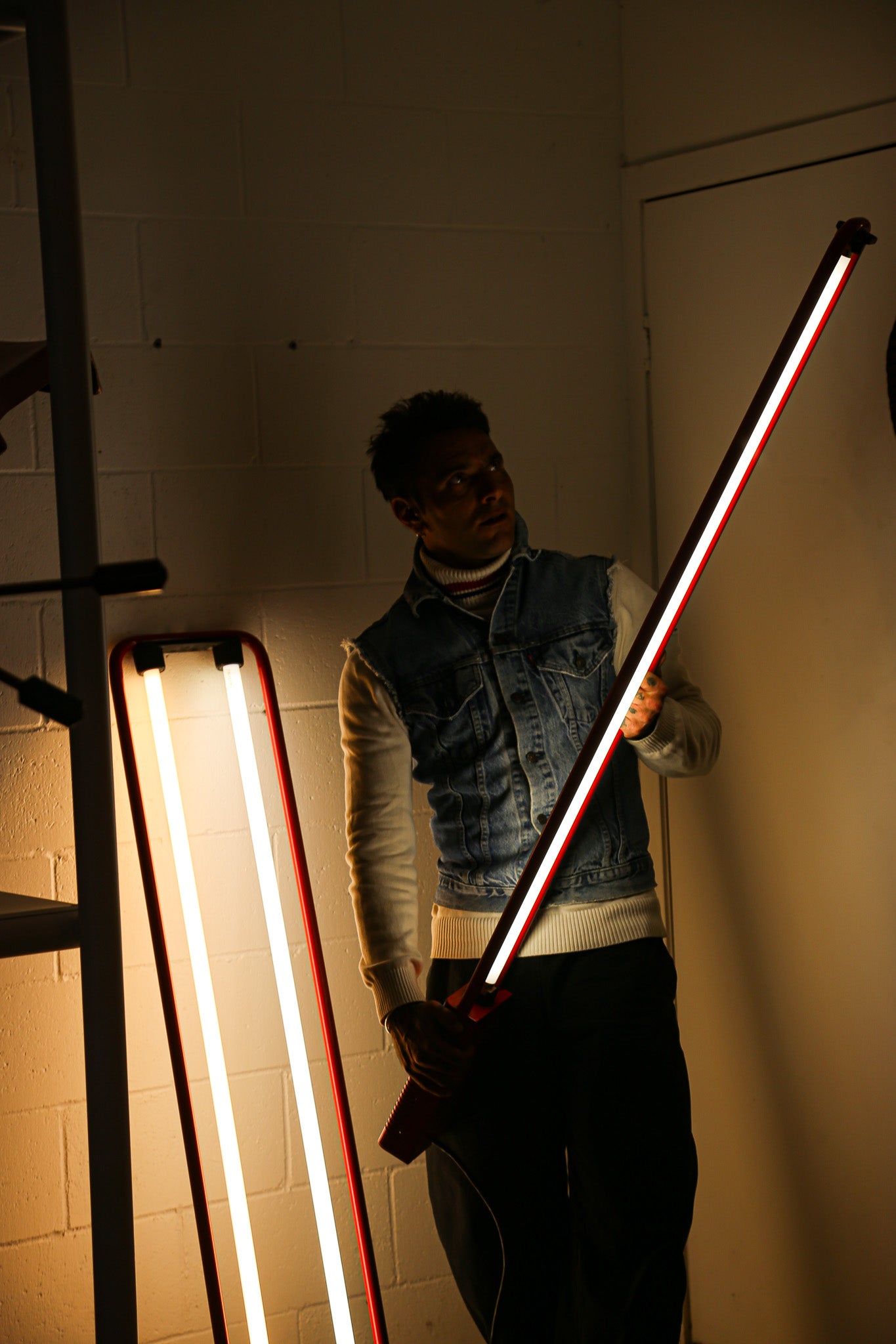 A person stands in a dimly lit room holding a long rectangular LED light bar that glows with white, red, and dark tones. Another similar light bar leans against the wall on the left.