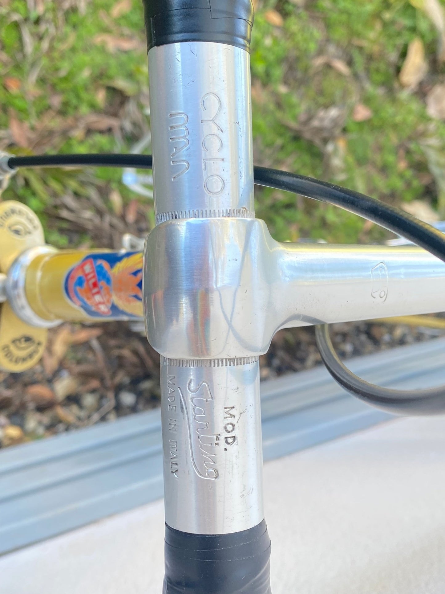 Close-up of a bicycle handlebar area showing a silver metal clamp with black grips and engraved text including 'Cyclo MXX' and 'Mod. Starling'.