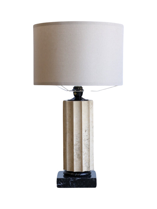 A table lamp with a base combining travertine and black marble, topped with a beige lampshade.