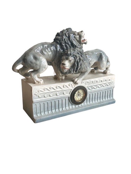 A Capodimonte sculpture featuring two lions with a clock integrated into the design, made of glazed porcelain with a grey and blue color palette.