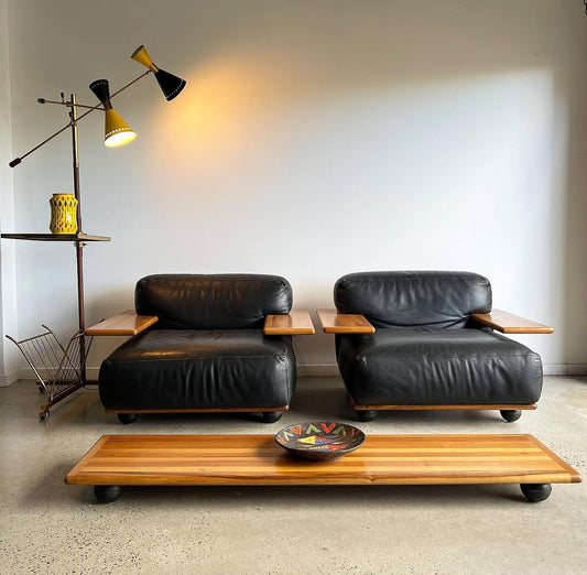 A pair of mid-century modern black leather armchairs with light wood side tables, positioned behind a matching low wooden coffee table in a minimalist room with a concrete floor and a warm wall lamp.