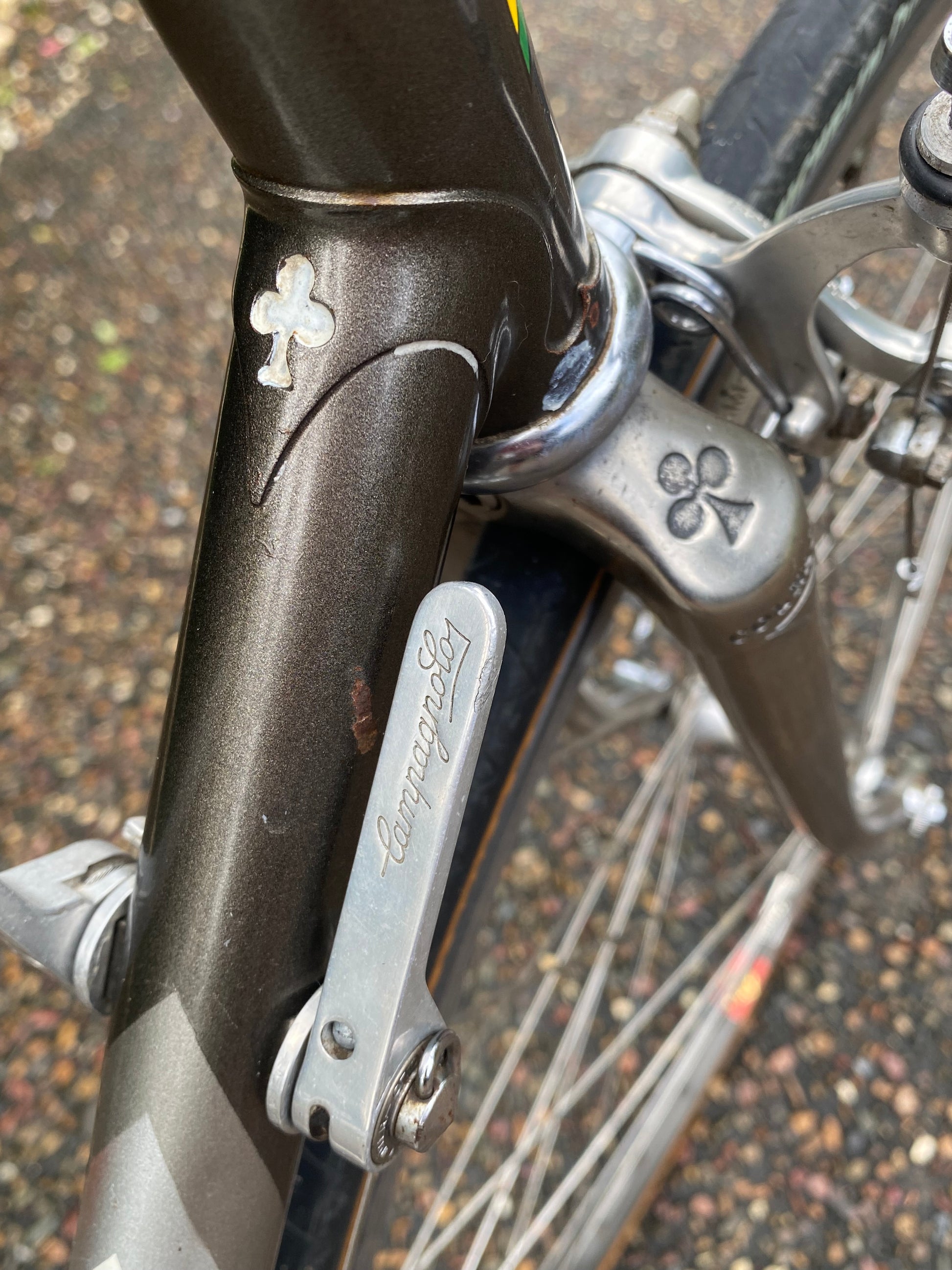 Close-up of a vintage bicycle frame and handlebar area, showing a dark metallic frame with silver metal components and a gravel ground background.