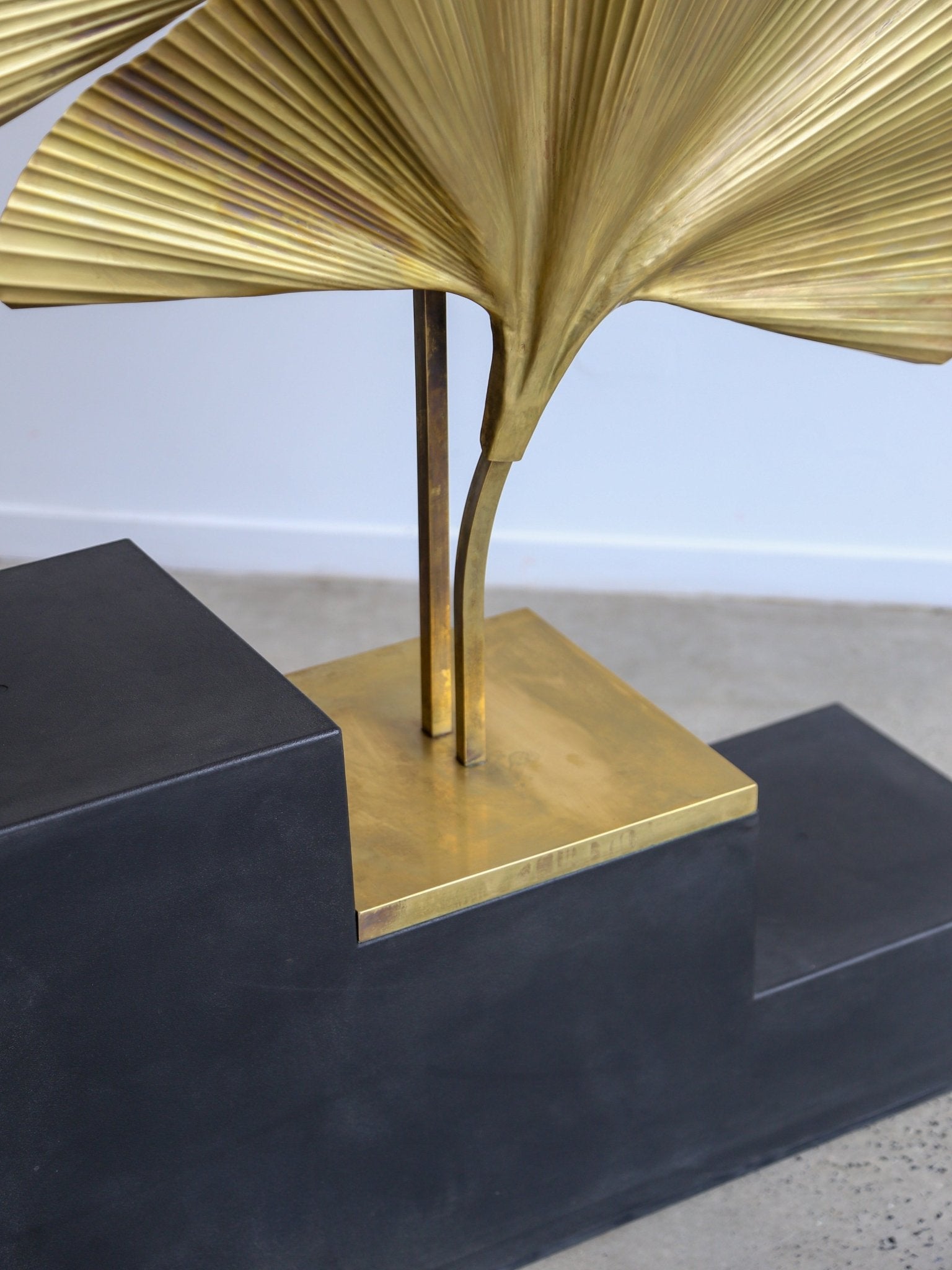 Gold leaf-like sculpture on a black pedestal against a light gray background
