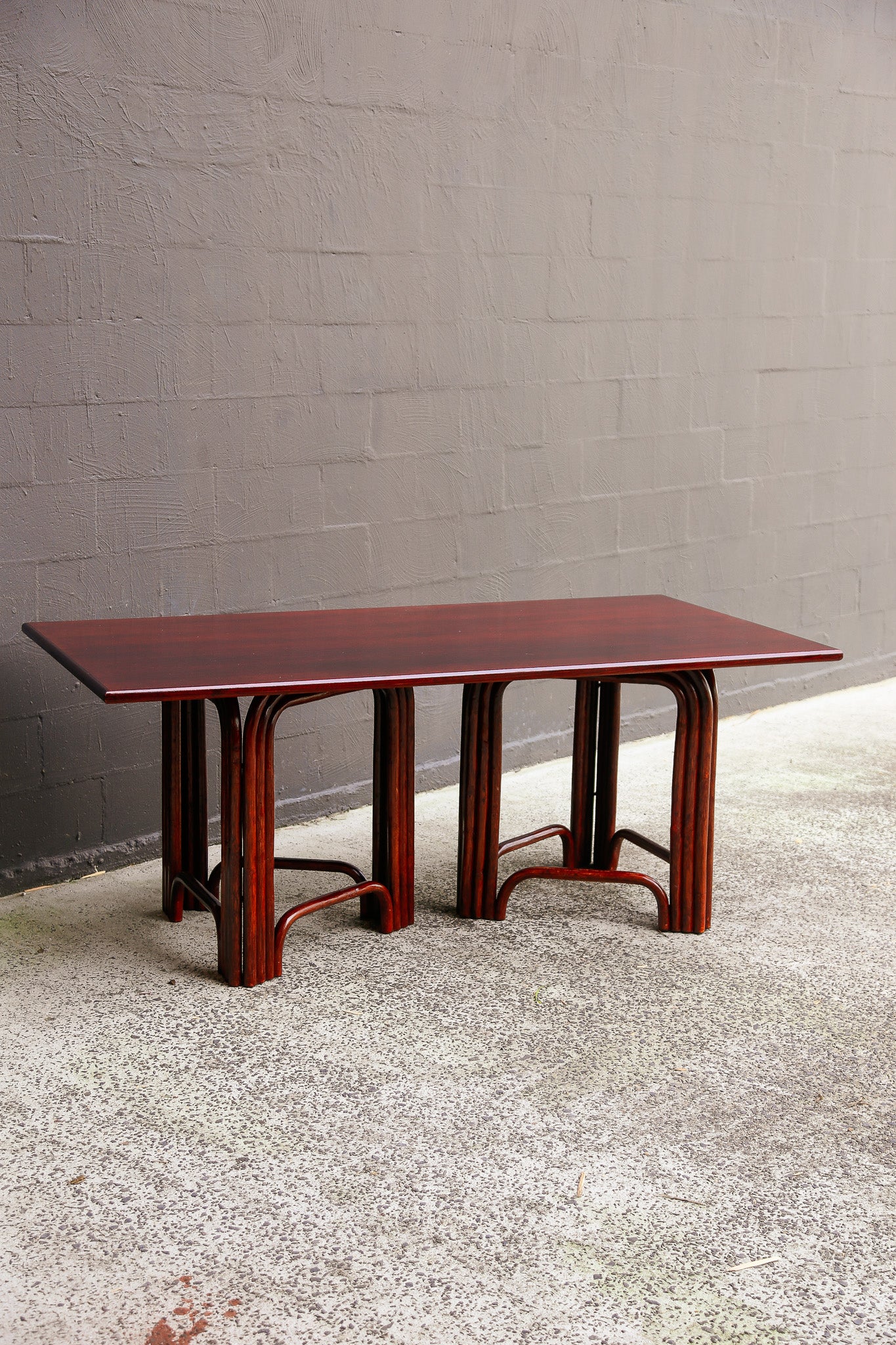 A rectangular wooden dining table with a glossy reddish-brown mahogany top and matching curved legs, set against a gray brick wall and concrete floor.