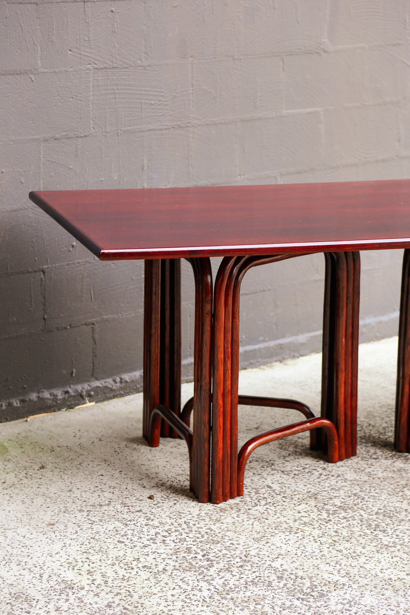 A mid-century modern wooden table with a reddish-brown look finish, featuring curved bentwood legs and a geometric top, set against a gray brick wall and concrete floor.
