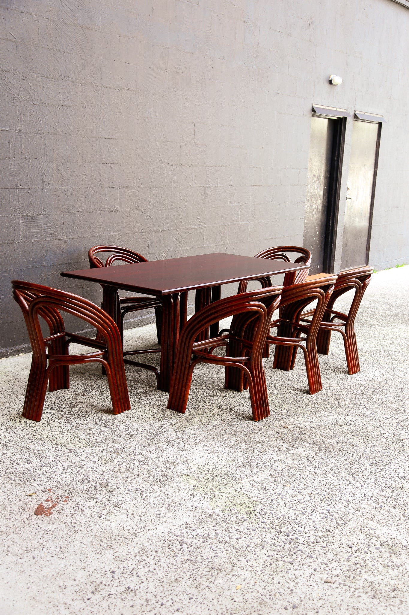 A dark reddish-brown wooden dining table with six matching curved-back chairs, set on a gravel surface against a gray concrete block wall.