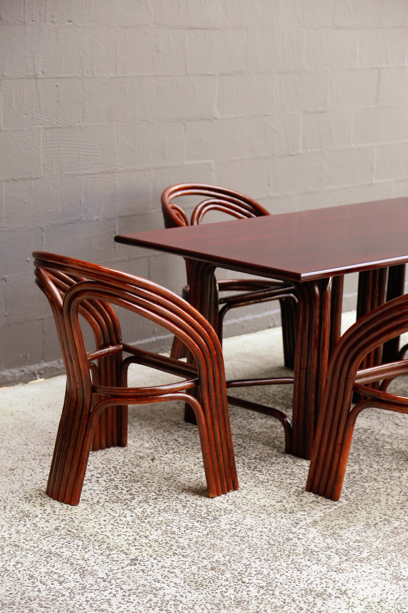 A mid-century modern wooden dining set consisting of a rectangular table and two matching curved armchairs with a rich reddish-brown finish, set against a gray brick wall and speckled concrete floor.