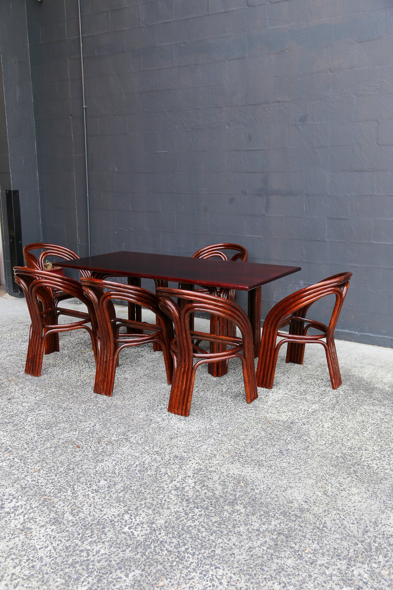A dark reddish-brown wooden dining table with six matching curved-back chairs, set against a gray concrete block wall and textured gray floor.