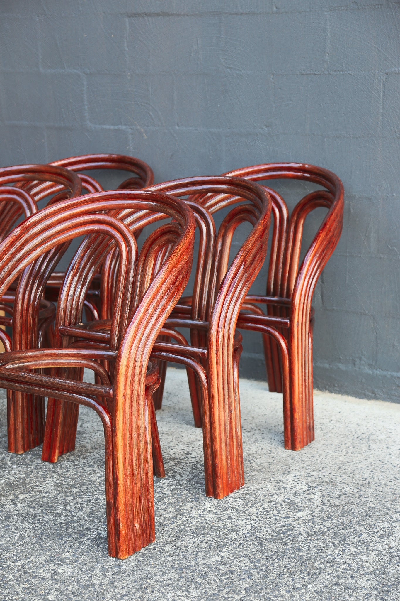 A group of bent bamboo chairs with a glossy reddish-brown finish, arranged against a gray painted brick wall on a concrete floor.