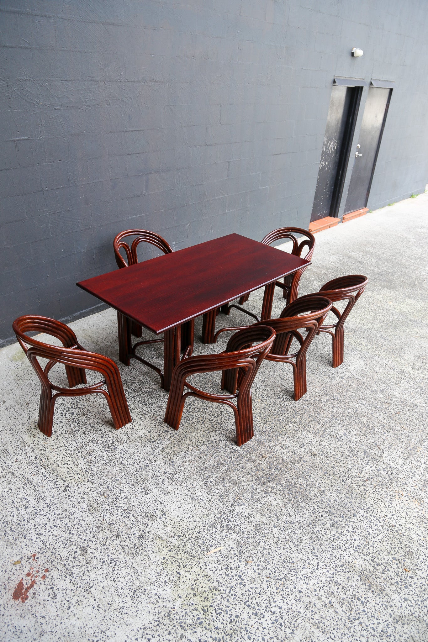 A dark reddish-brown wooden dining table set with six matching curved-back chairs, placed on a speckled concrete floor against a dark gray brick wall.