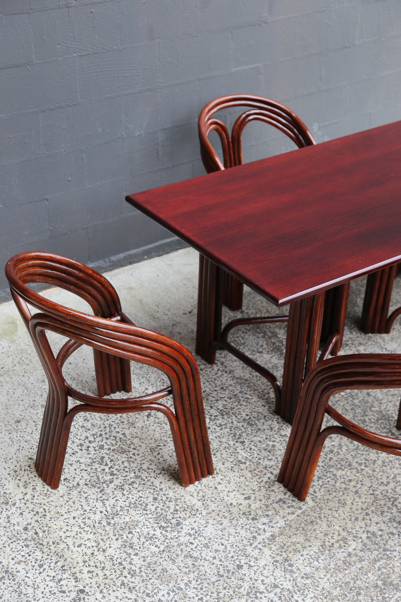 A dark reddish-brown wooden dining table with matching curved wooden chairs, set against a gray brick wall and speckled concrete floor.