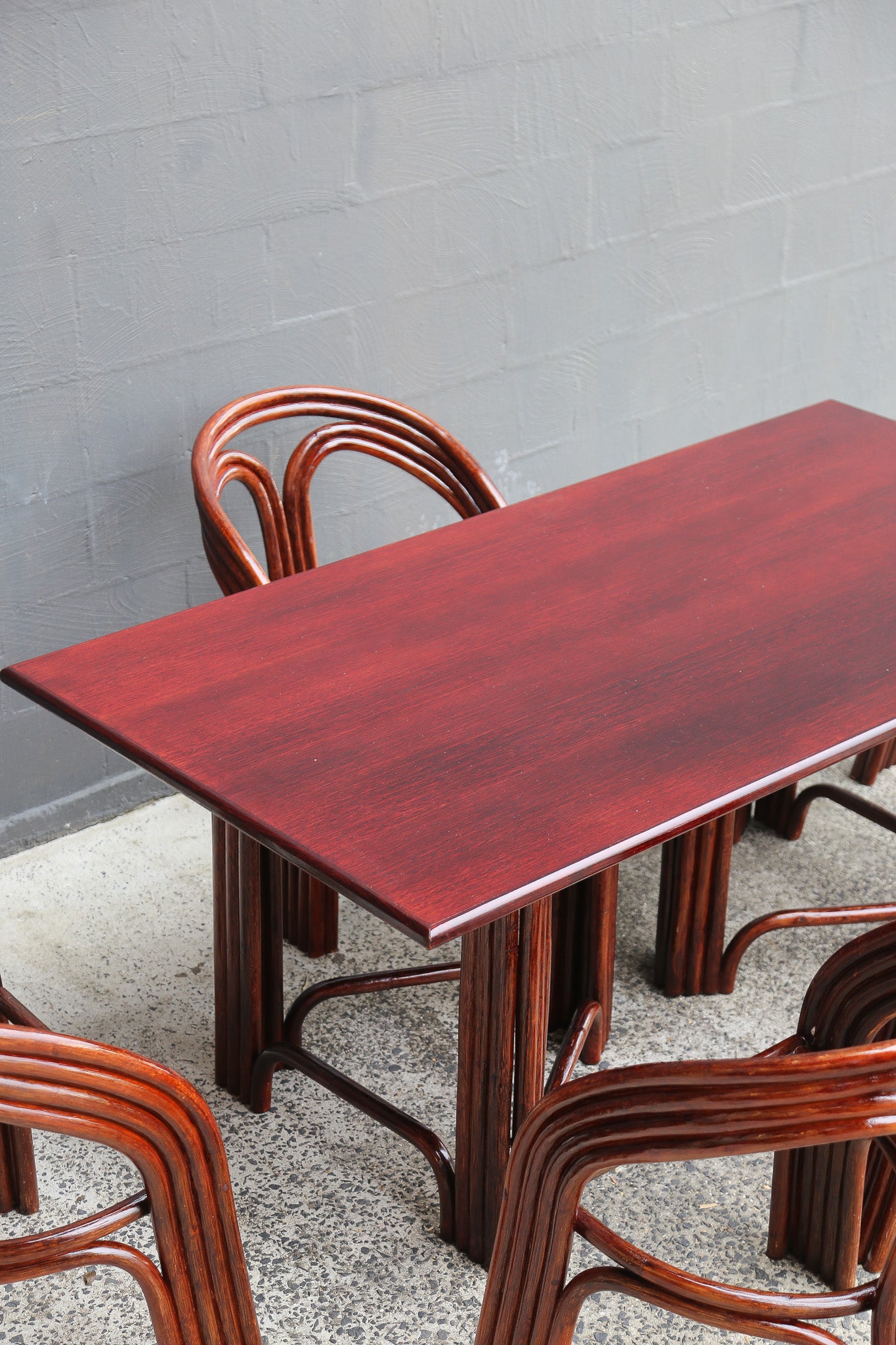 A rectangular dining table with a reddish-brown wood grain top and matching curved wooden chairs, set against a gray brick wall and speckled concrete floor.
