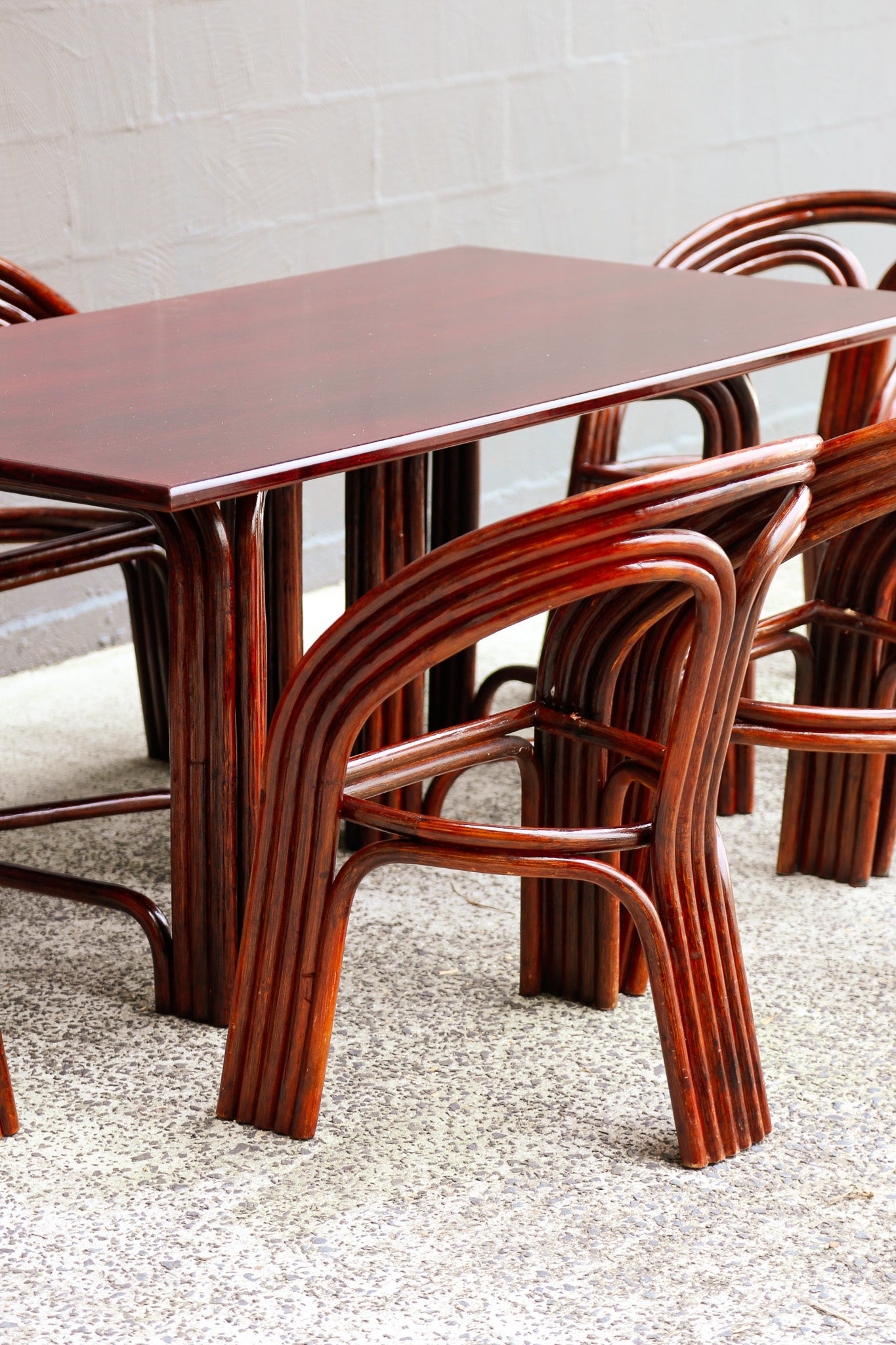 A dark reddish-brown bamboo dining table with matching curved wooden chairs, set on a speckled gray floor against a light gray brick wall.