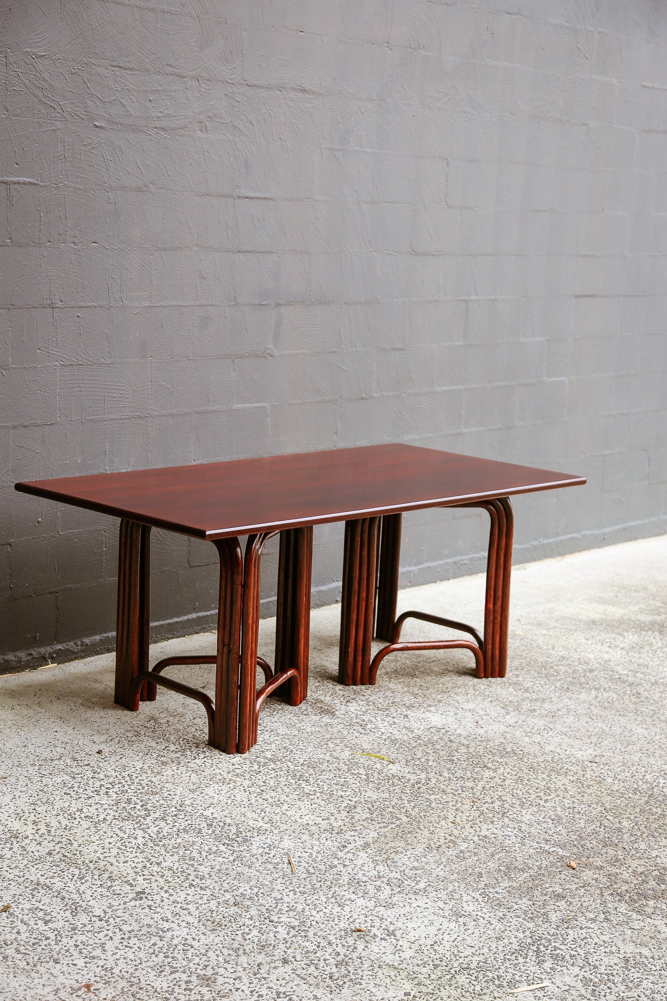 A rectangular wooden dining table with a reddish-brown finish and curved bent bamboo legs, set against a gray brick wall and concrete floor.