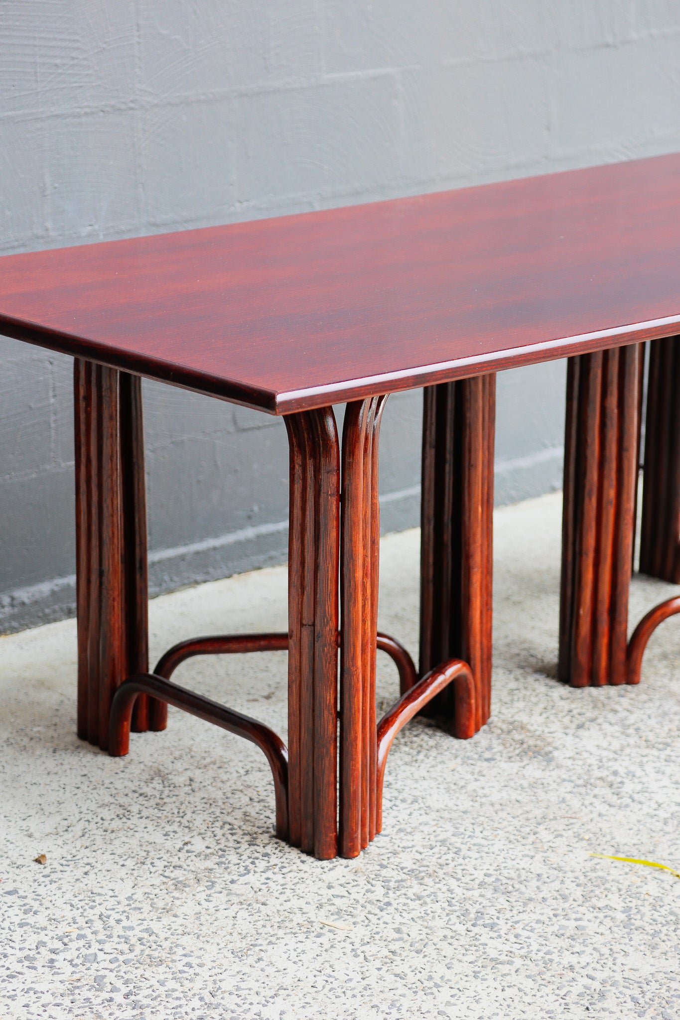 A rectangular wooden dining table with a glossy reddish-brown mahogany finish, featuring fluted legs and curved aprons, set against a gray concrete wall and floor.