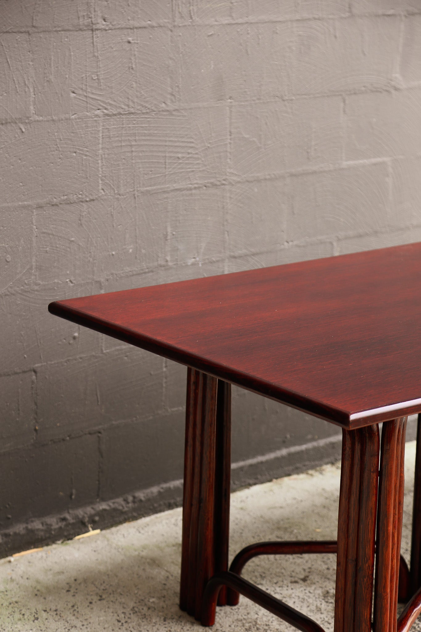 A close-up of a dark reddish-brown wooden table corner with a smooth top and slatted legs, set against a gray painted brick wall and concrete floor.