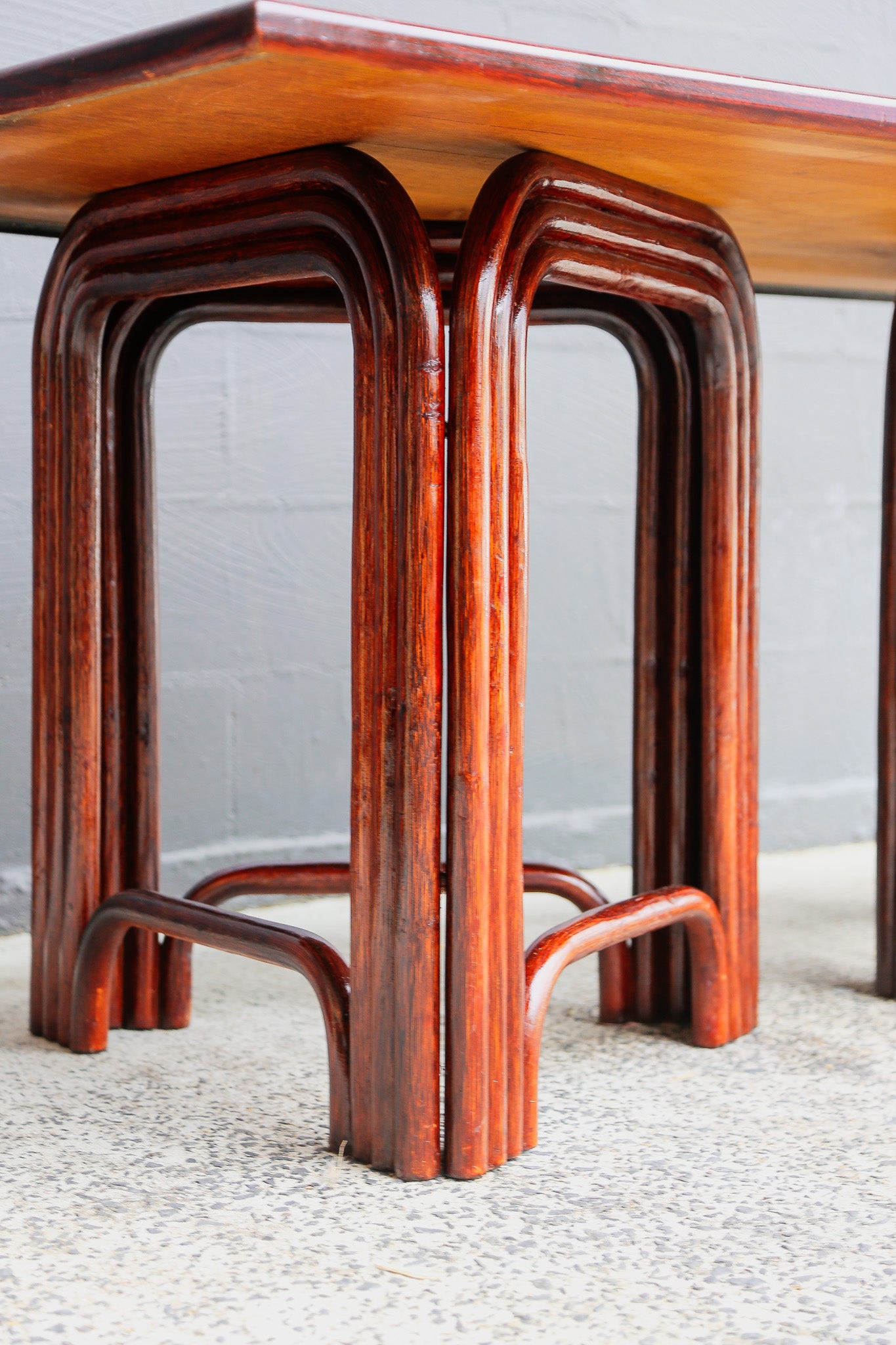 A close-up of a reddish-brown wooden table leg featuring a curved, multi-layered bentwood design, set against a gray wall and speckled floor.