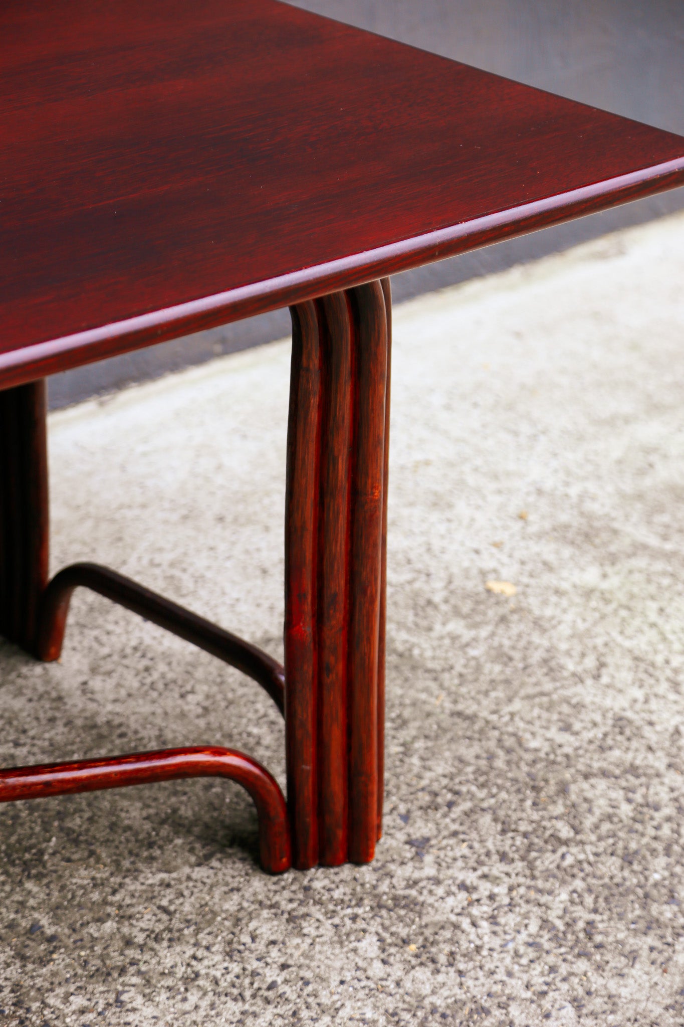 A close-up of a dark reddish-brown wooden table with a glossy finish, featuring curved bentwood legs resting on a concrete floor.