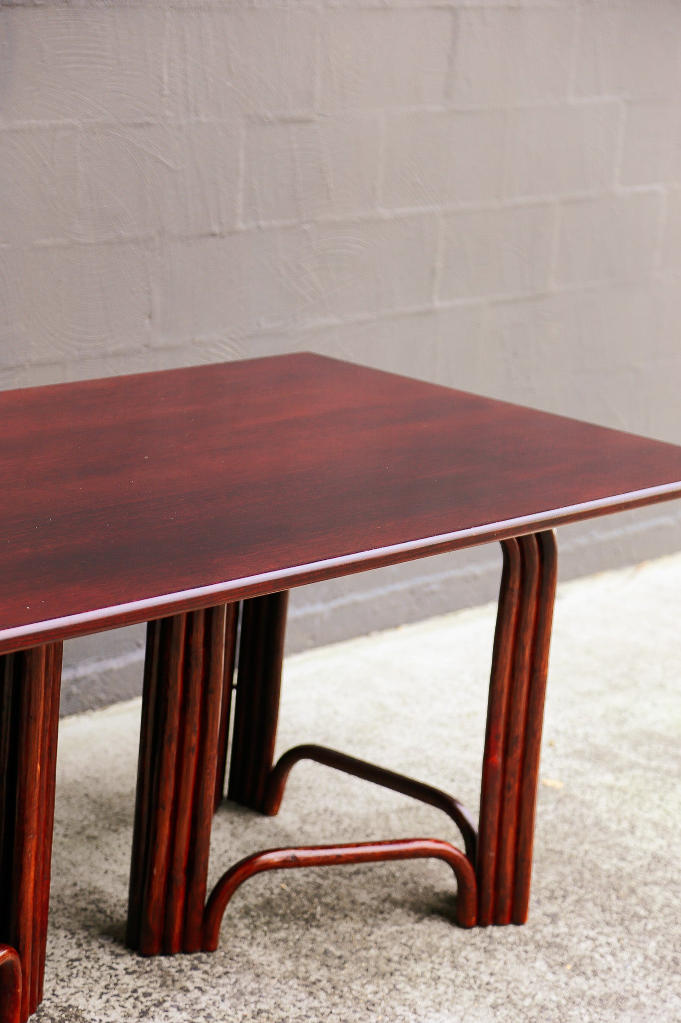 A rectangular nesting side table with a glossy reddish-brown wood finish and curved metal support legs, shown against a gray brick wall and concrete floor.