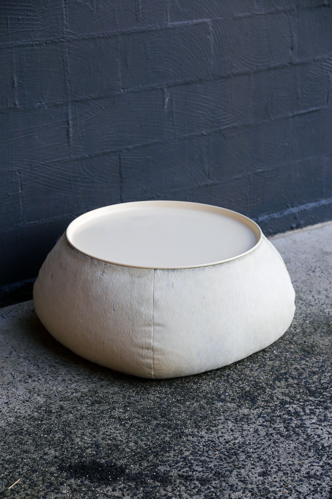 Beige fabric ottoman on a dark concrete floor against a dark blue brick wall.