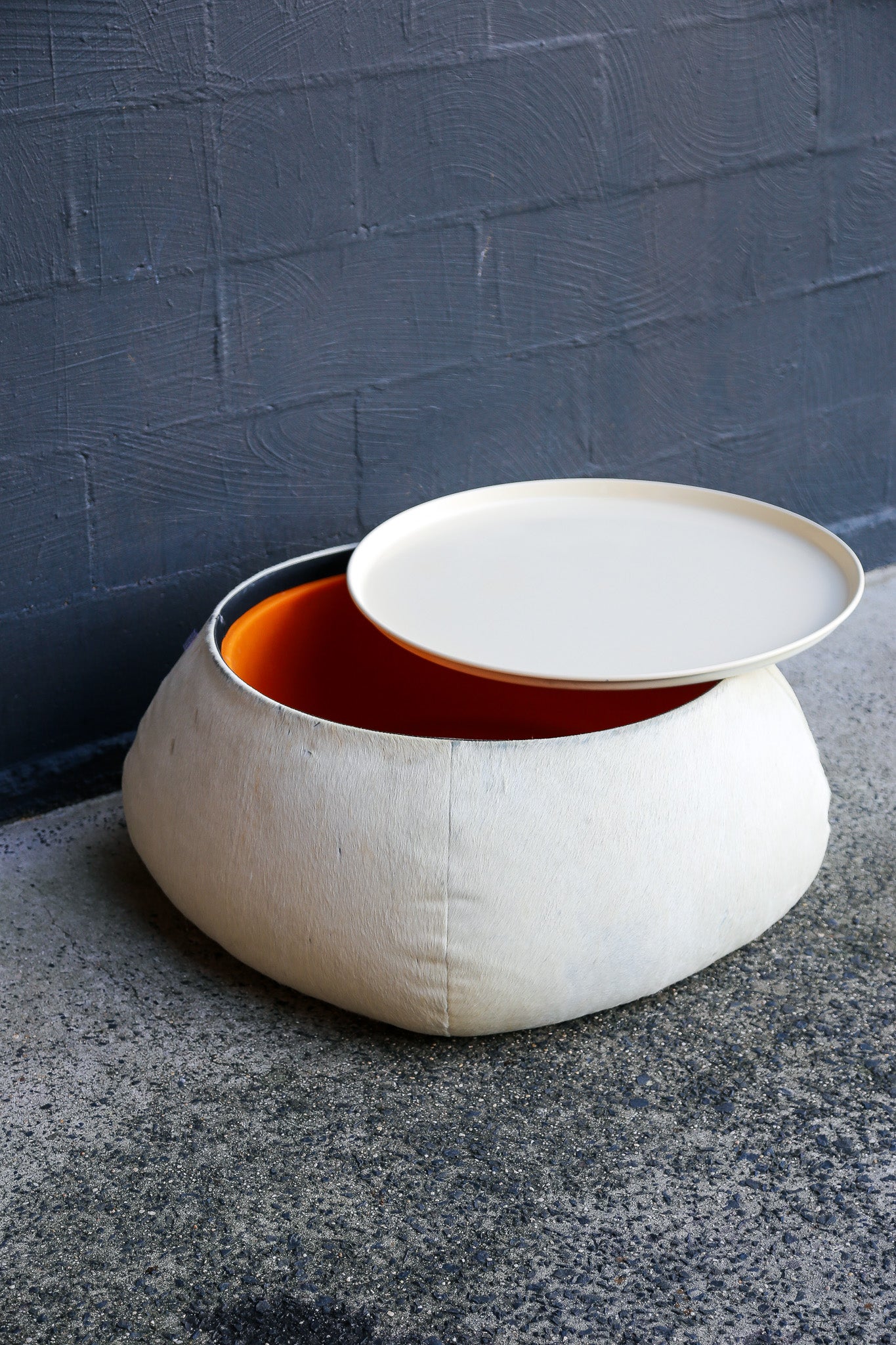 White ottoman with a lid on a gray surface against a dark brick wall.