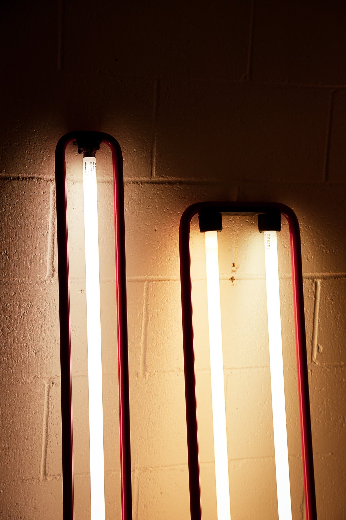 Two vintage vertical fluorescent light tubes with dark red frames mounted on a beige brick wall, casting a warm yellow glow.