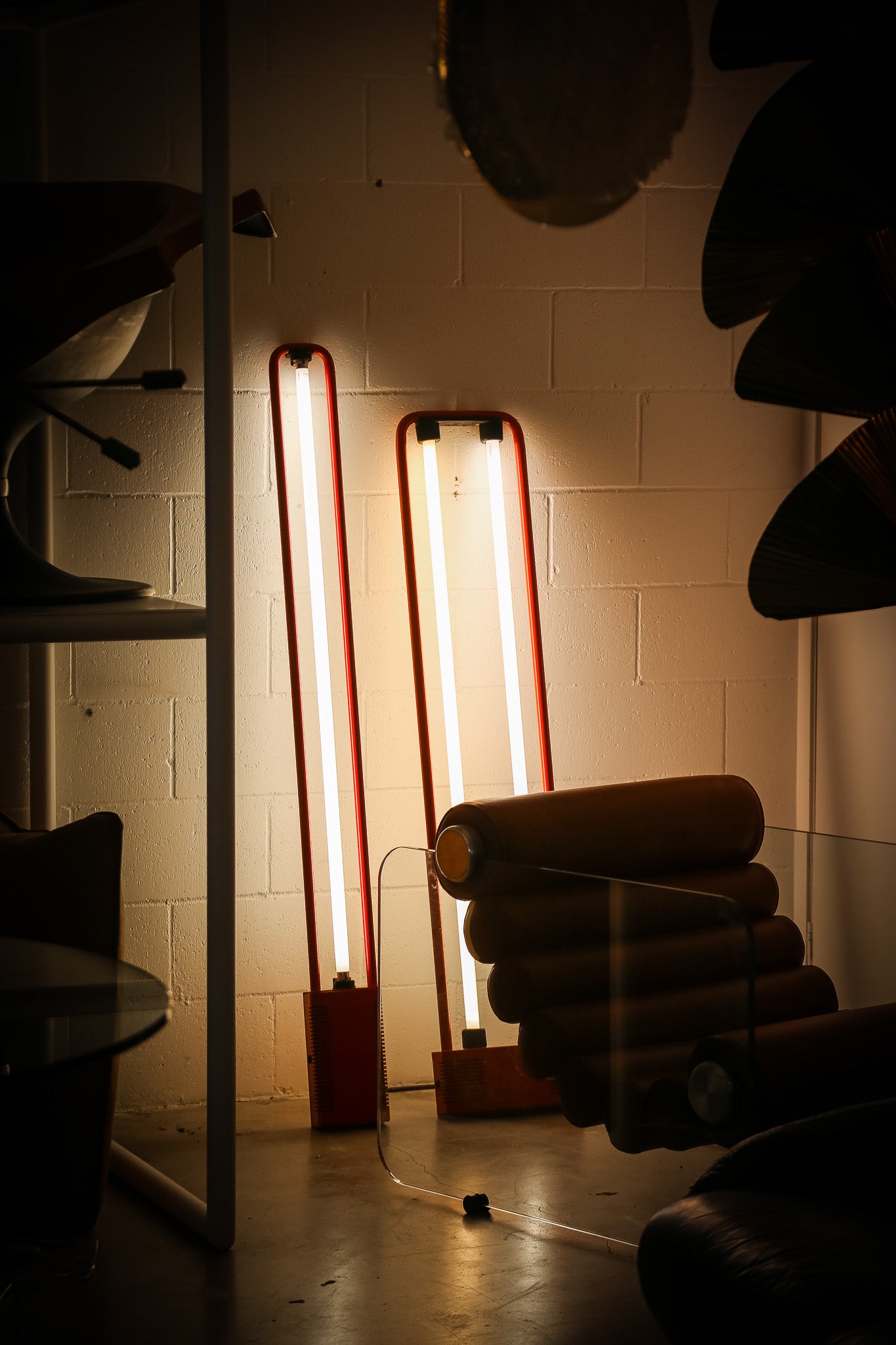 Two tall rectangular floor lamps with red metal frames and glowing white tubes, standing in a dimly lit room with a cinderblock wall and furniture partially visible.