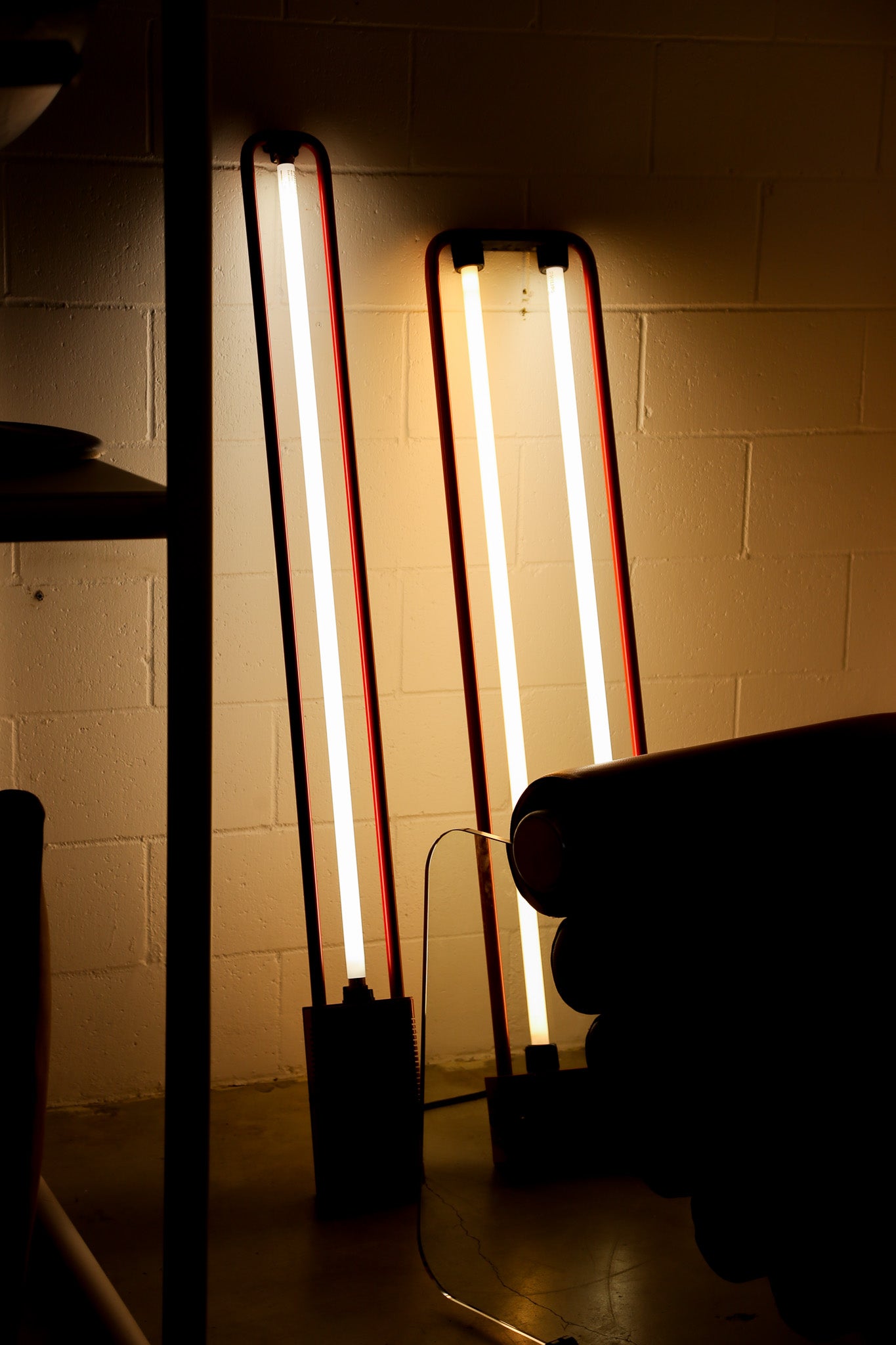 Two tall rectangular floor lamps with glowing vertical tubes and dark metal frames, standing against a beige brick wall in a dim interior.