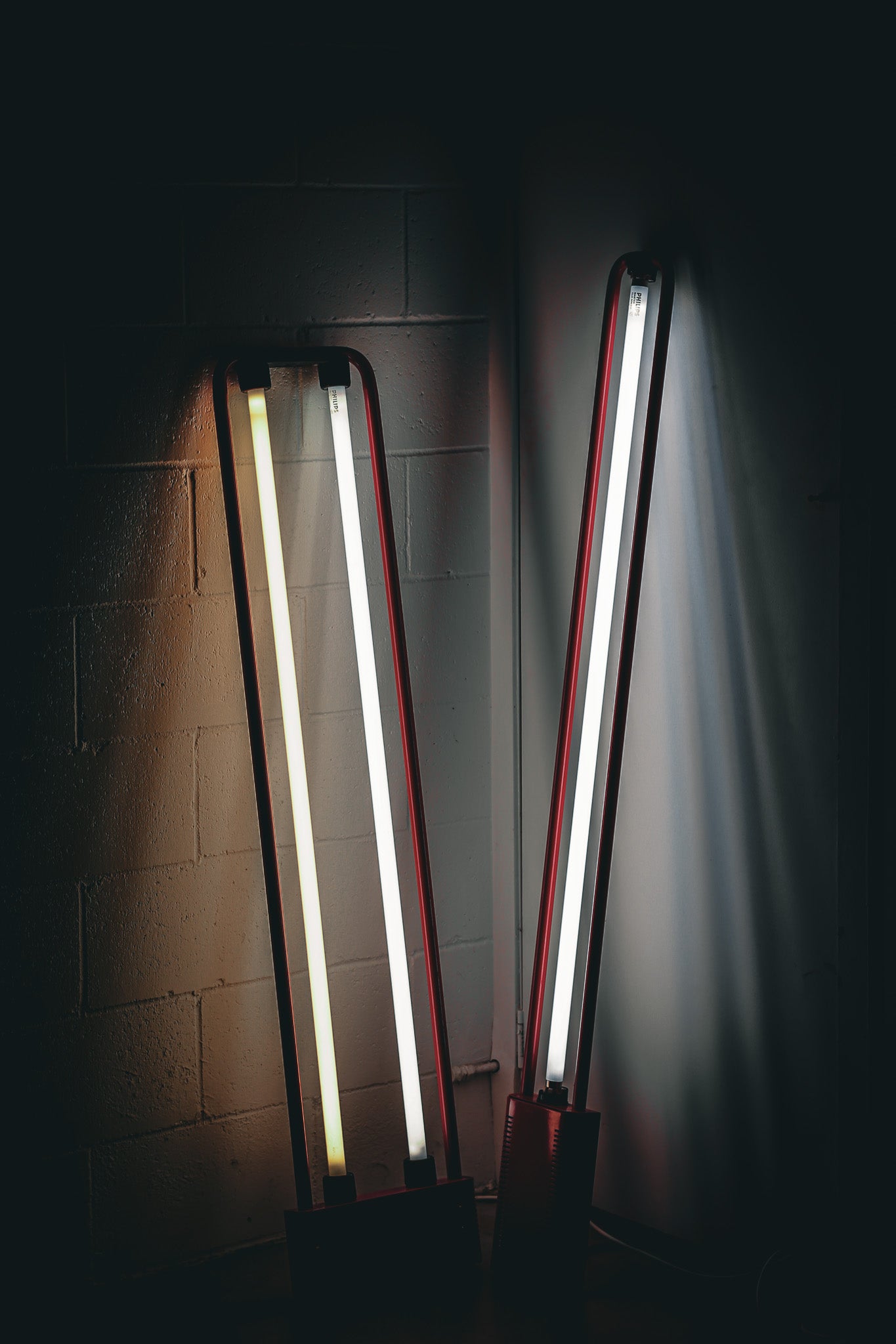 Two tall, narrow fluorescent tube light fixtures with red side rails, glowing brightly against a dark wall with a cinderblock texture.