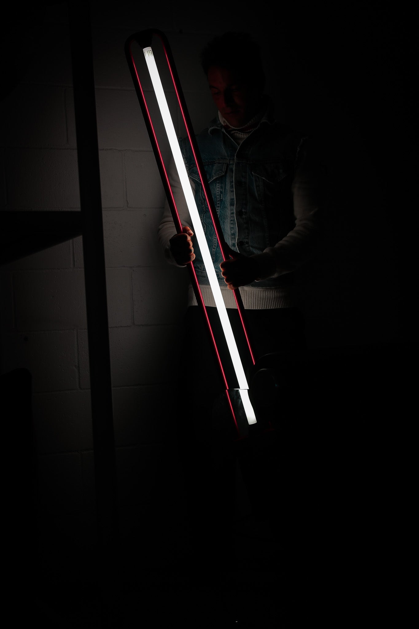 A long, narrow LED light bar with a bright white center and red outer edges, held by a person in a very dark room against a dim wall.