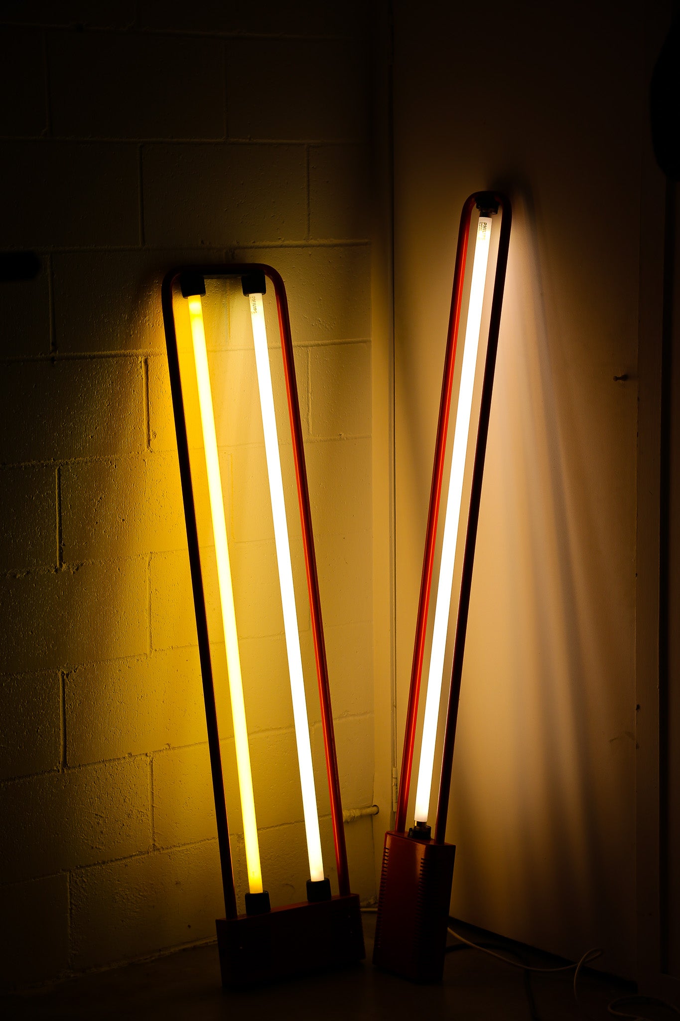 Two tall, narrow LED neon-style wall lights with glowing yellow and white tubes, mounted against a dark wall in a dim interior.
