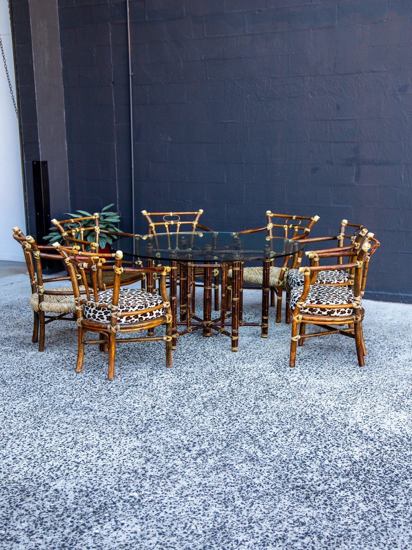 Set of bamboo chairs with patterned cushions around a glass dining table against a dark wall.