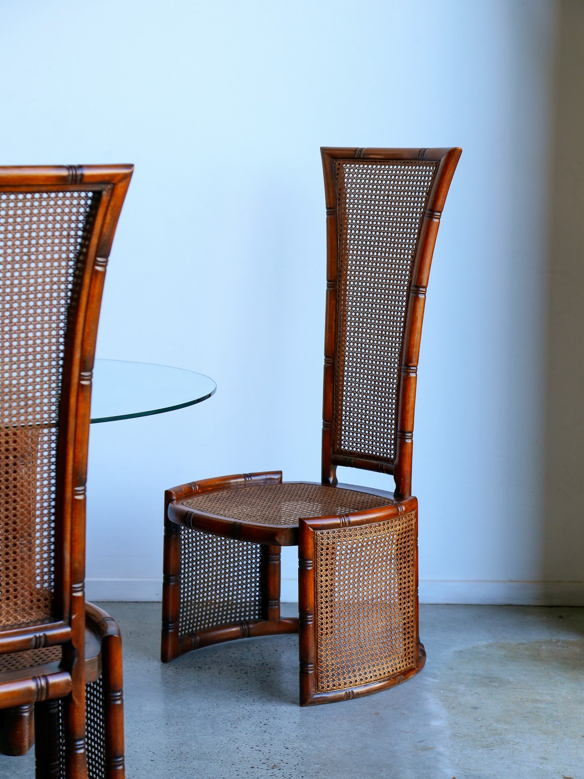 Two wooden chairs with wicker backrests and seat in a room with a plain wall and floor.