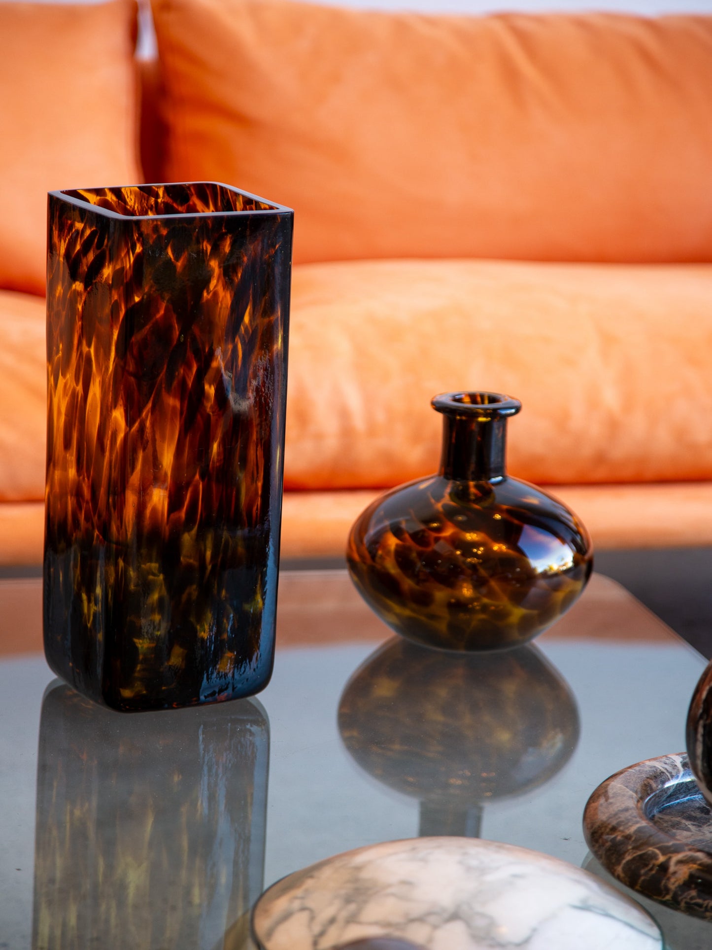 Italian vintage Murano-style tortoiseshell glass vase and rounded bottle vase displayed on a glass coffee table with marble decor, against orange velvet sofa