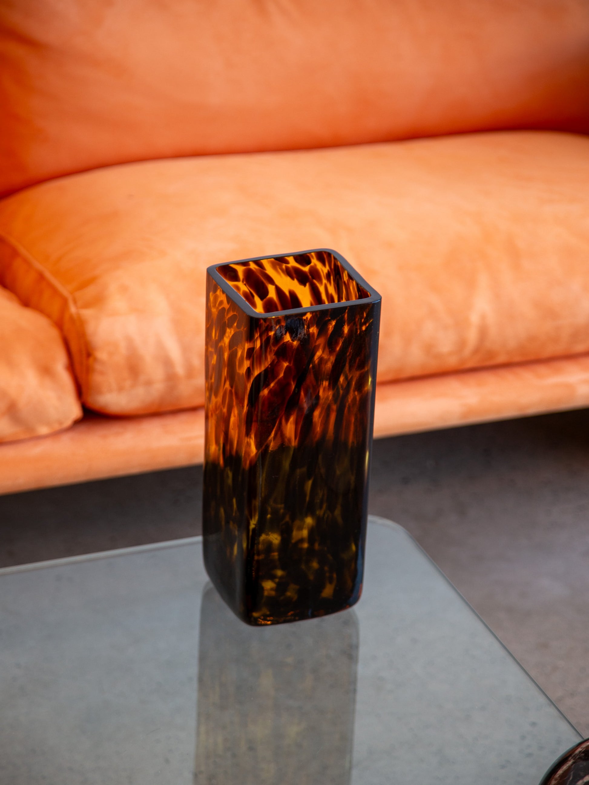 Rectangular tortoiseshell Murano-style glass vase in amber and black, styled on a glass table with orange sofa in the background