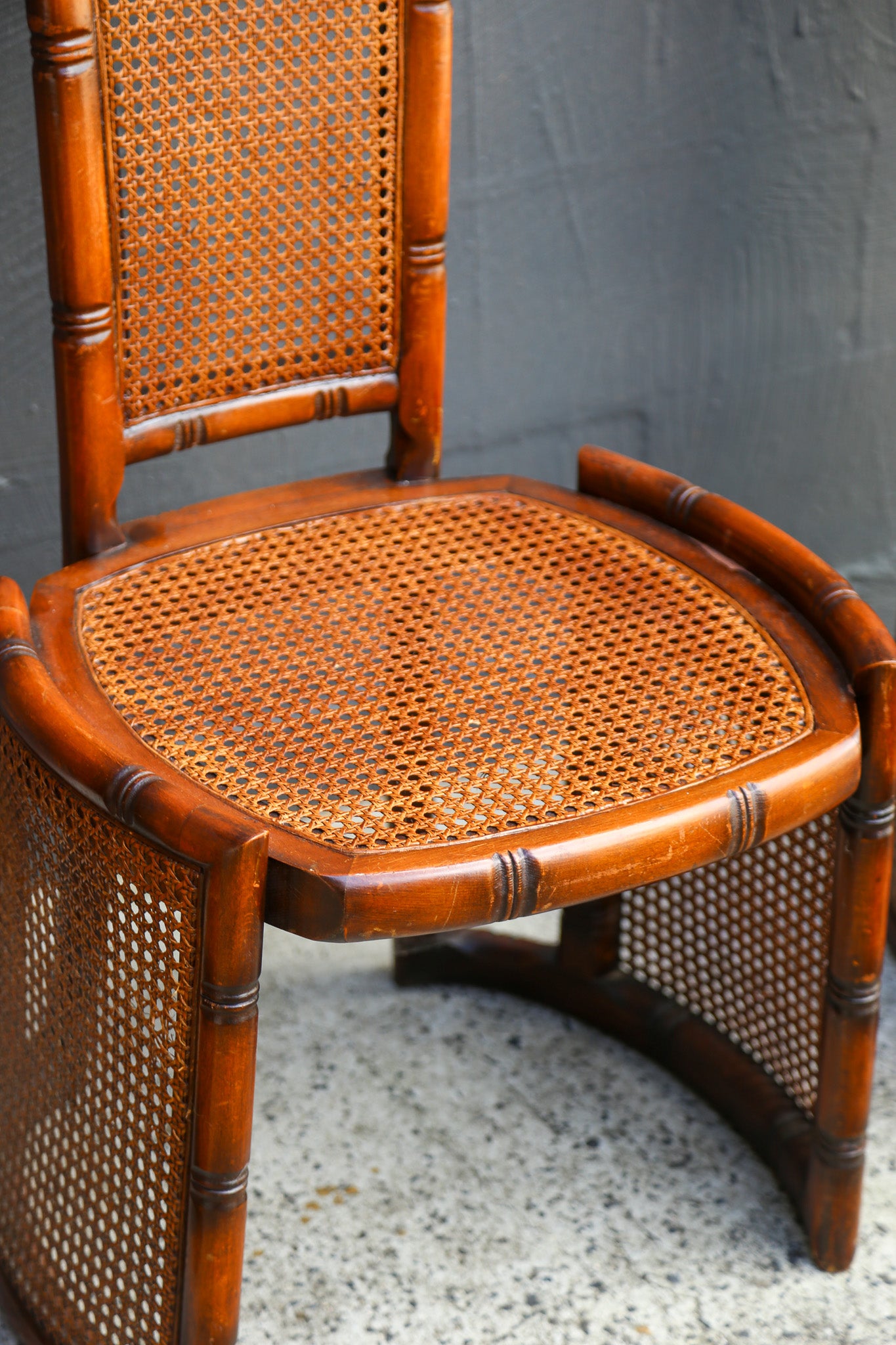 A curved wooden dining chair with a woven cane seat and backrest, shown against a gray wall and speckled floor.