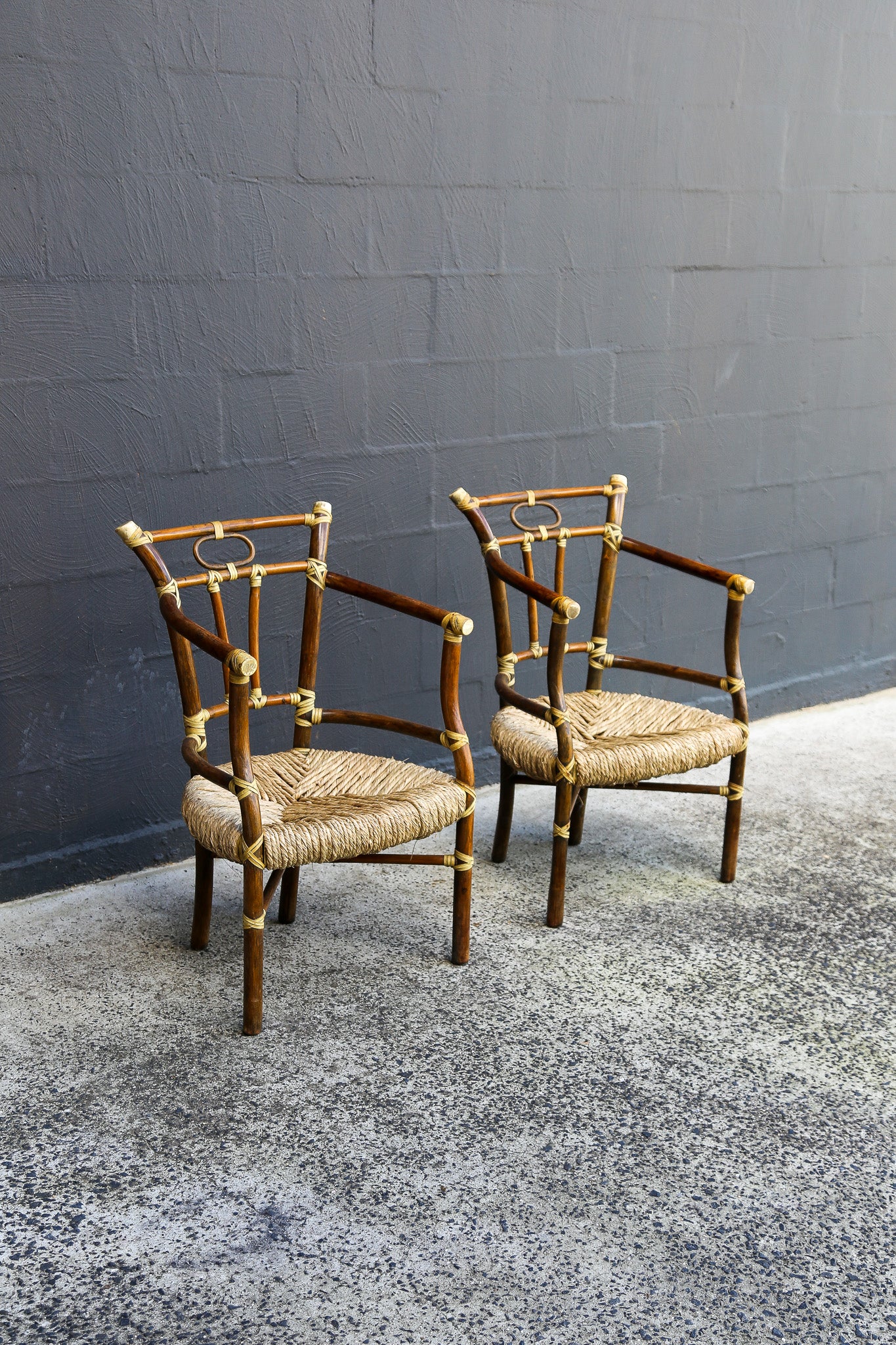 Two vintage-style bamboo armchairs with woven straw seats, positioned on a speckled concrete floor against a dark gray brick wall.