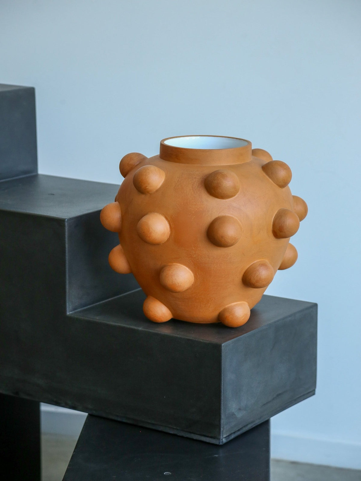Ceramic vase with a textured surface on a black pedestal against a light blue wall.