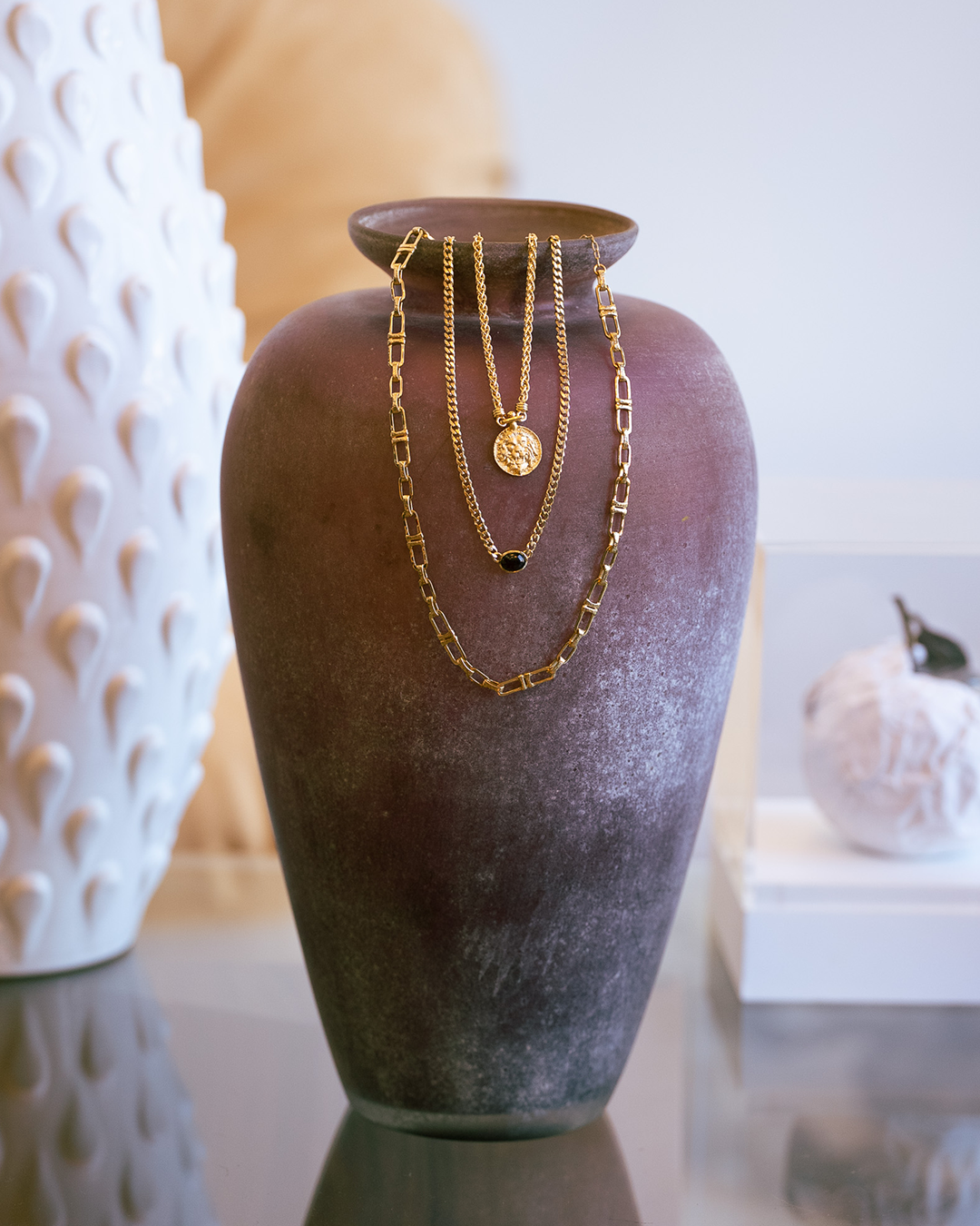 Three gold necklaces on a textured brown vase with a blurred indoor setting.