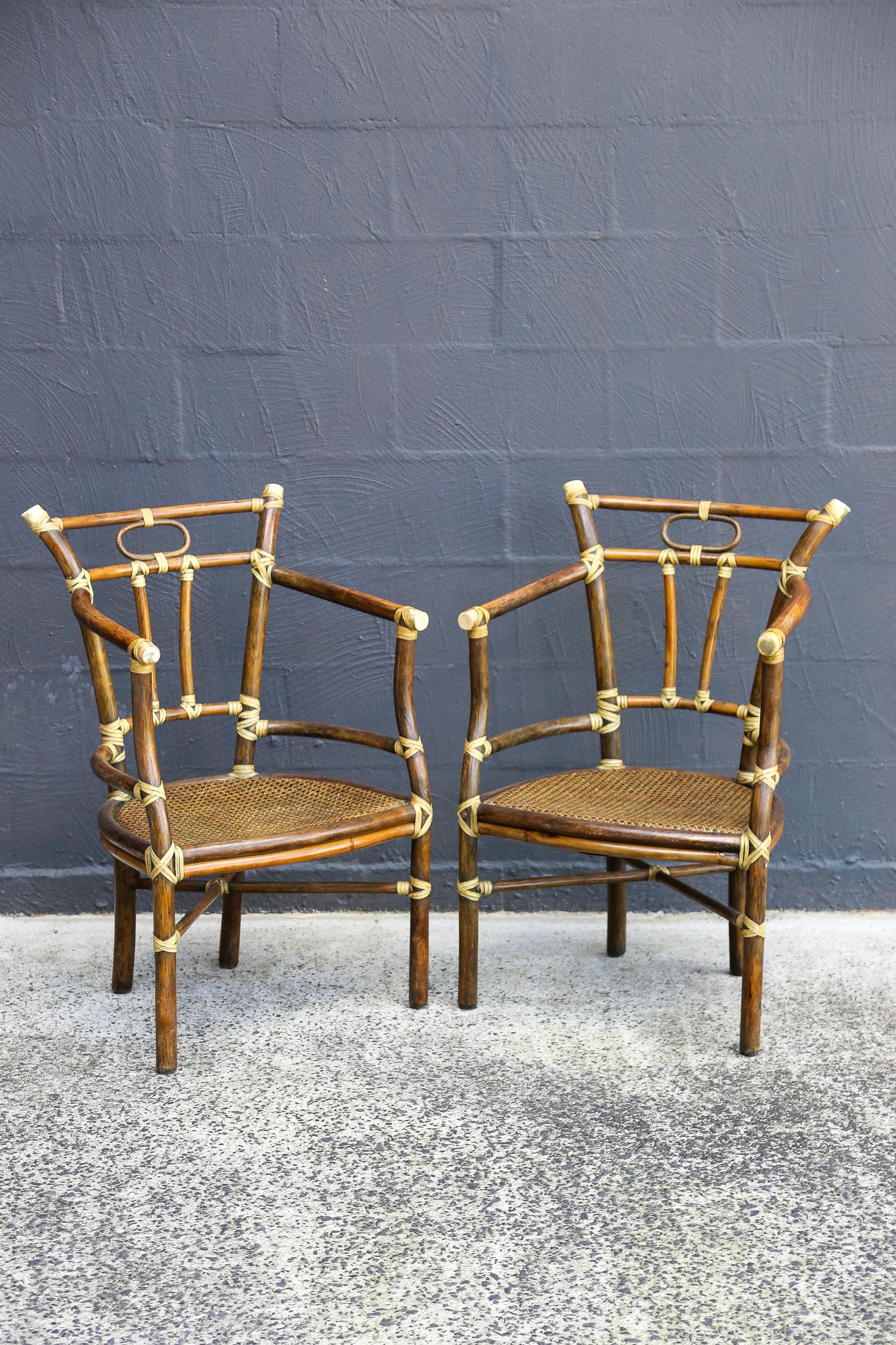 Two matching bamboo armchairs with woven cane seats and brass hardware, set against a dark gray brick wall and concrete floor.
