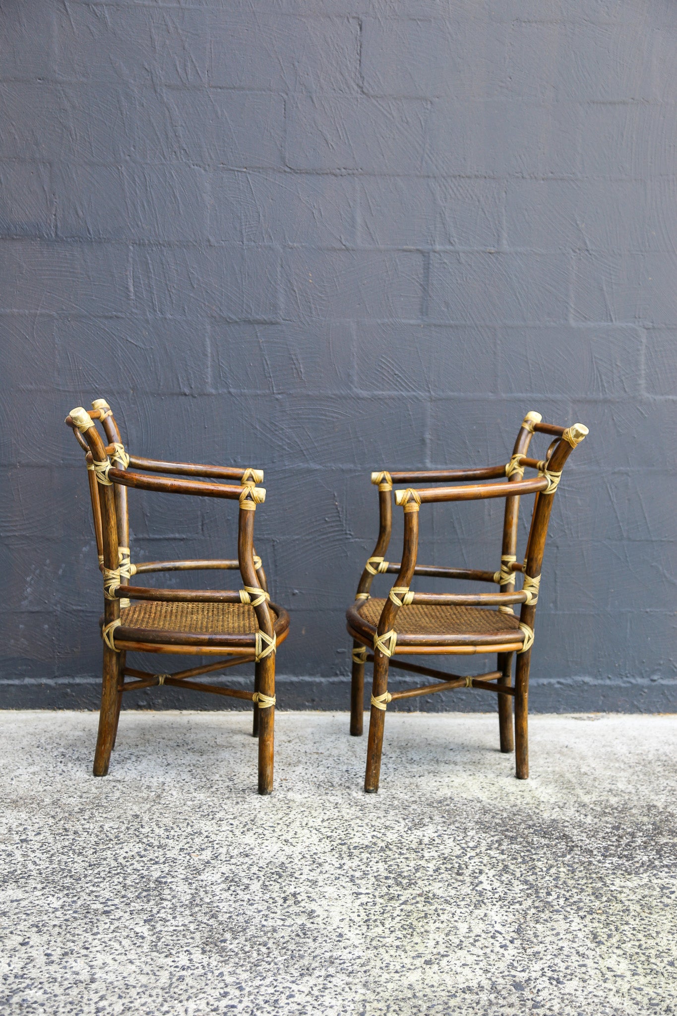Two vintage-style bamboo armchairs with woven cane seats and brass hardware, positioned side-by-side against a dark gray brick wall.