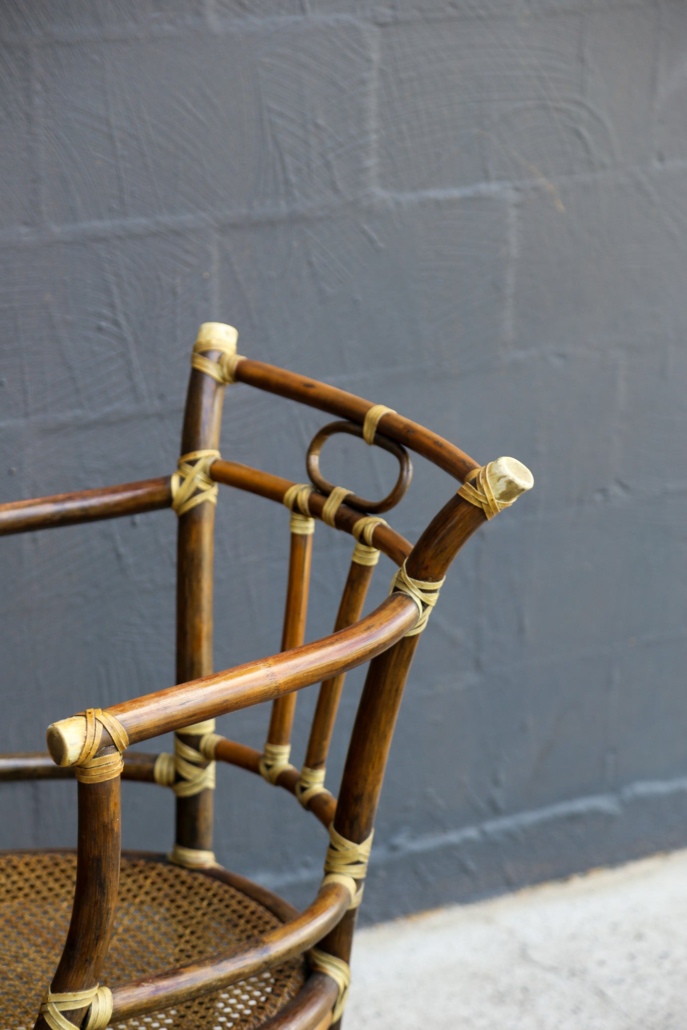 A close-up of a rustic woven rattan chair with curved brown wooden arms and backrest, tied together with natural tan rope, set against a dark gray painted brick wall.