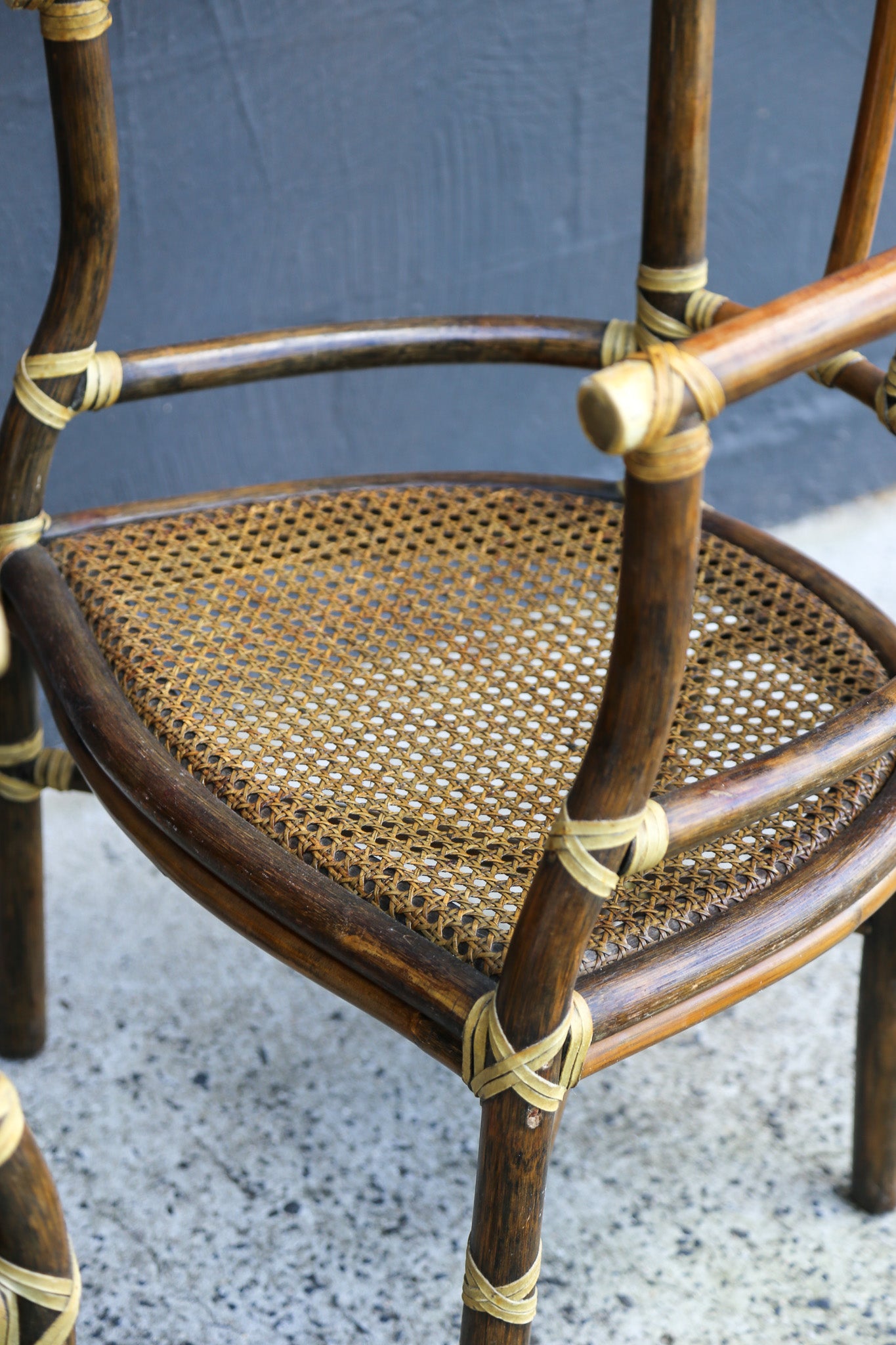 A close-up of a rustic wooden chair with a woven cane seat and light tan rope binding, set against a dark gray wall and speckled gray floor.