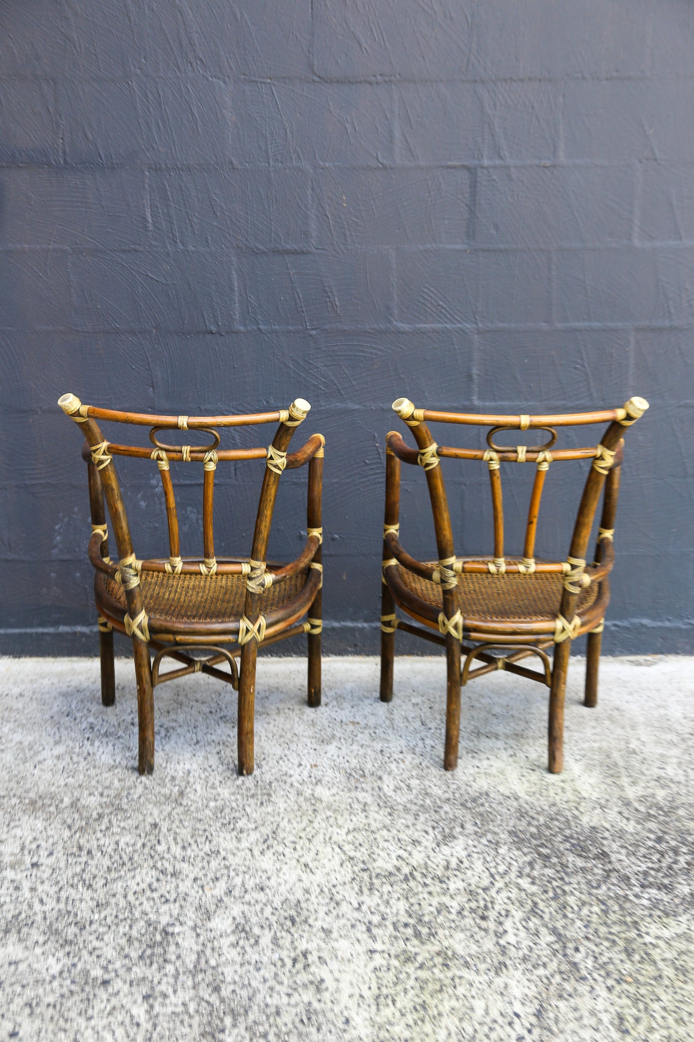 Two matching mid-century modern bamboo armchairs with woven cane seats and brass accents, positioned side-by-side against a dark gray brick wall.
