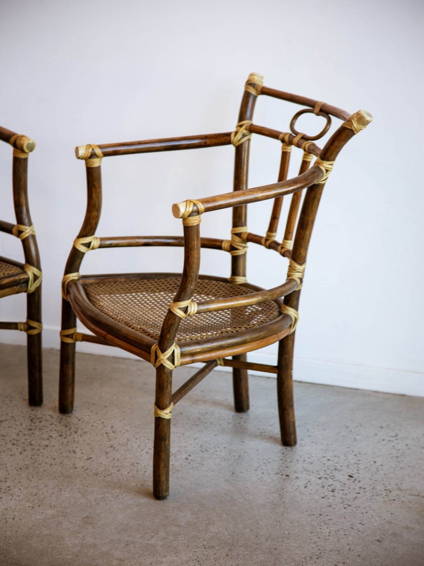 A mid-century modern bamboo armchair with a woven cane seat, positioned in a minimalist room with a concrete floor and white wall. Another matching chair is partially visible on the left.