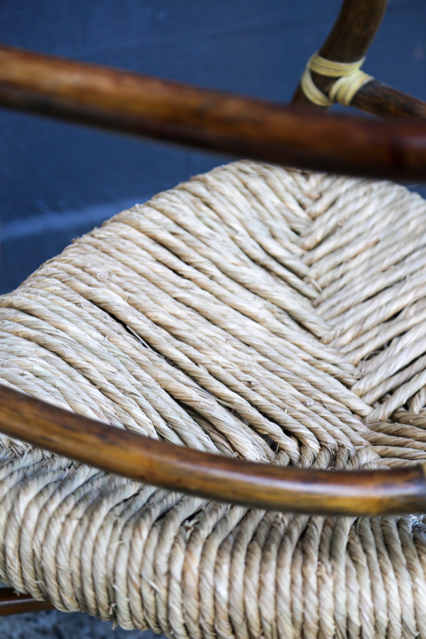 Close-up of a woven natural fiber chair seat with curved dark brown wooden armrests and a matching wrapped leg, set against a dark blue background.