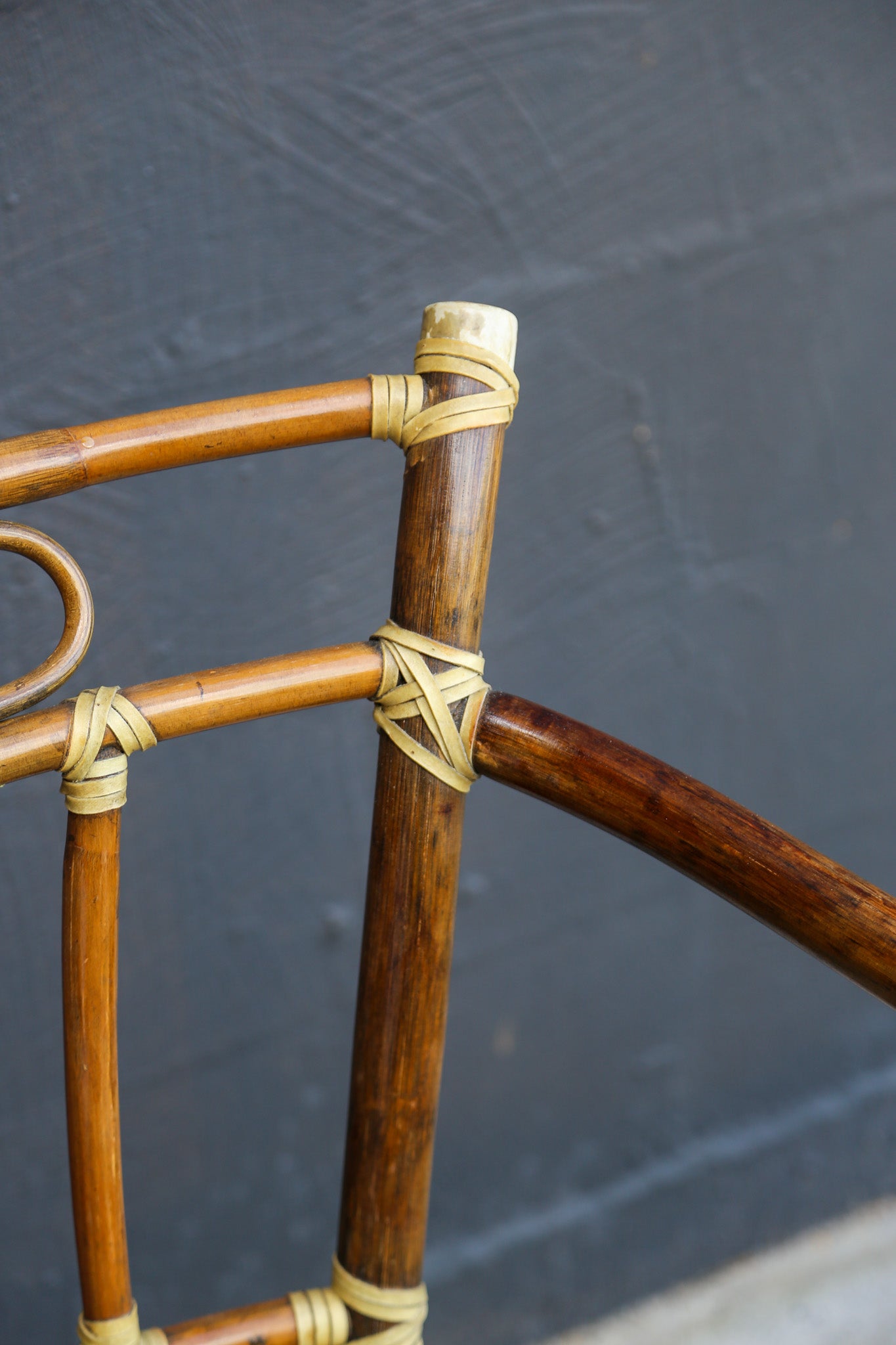 Close-up of a wooden chair frame made of dark brown bamboo or rattan tubes, bound together with light tan natural fiber wraps against a dark gray textured background.