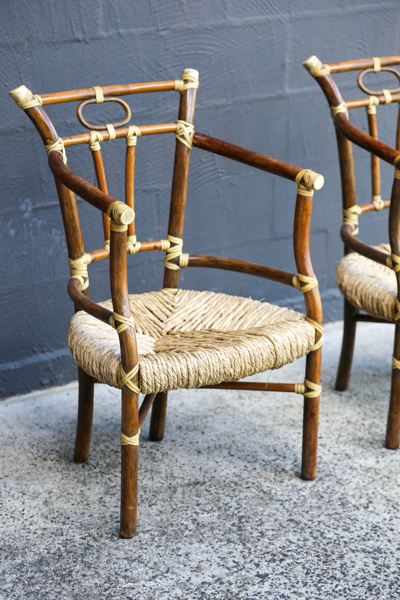 A mid-century modern armchair made of curved brown bamboo or rattan tubes with a woven natural fiber seat, set against a dark gray textured wall.