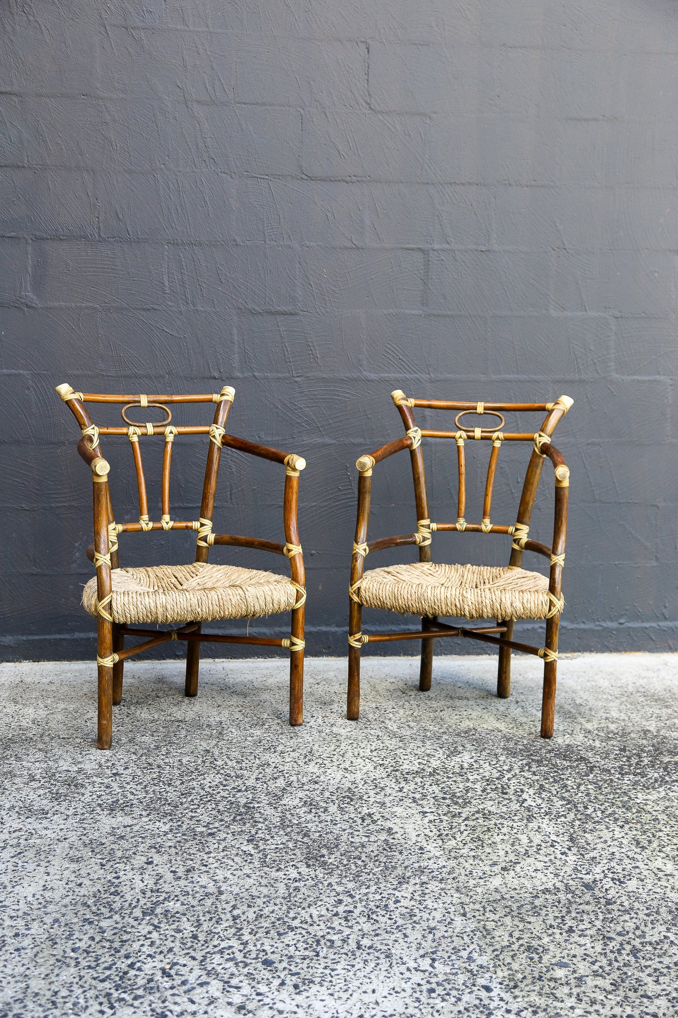 Two matching bamboo armchairs with woven straw seats, positioned side-by-side against a dark gray brick wall.